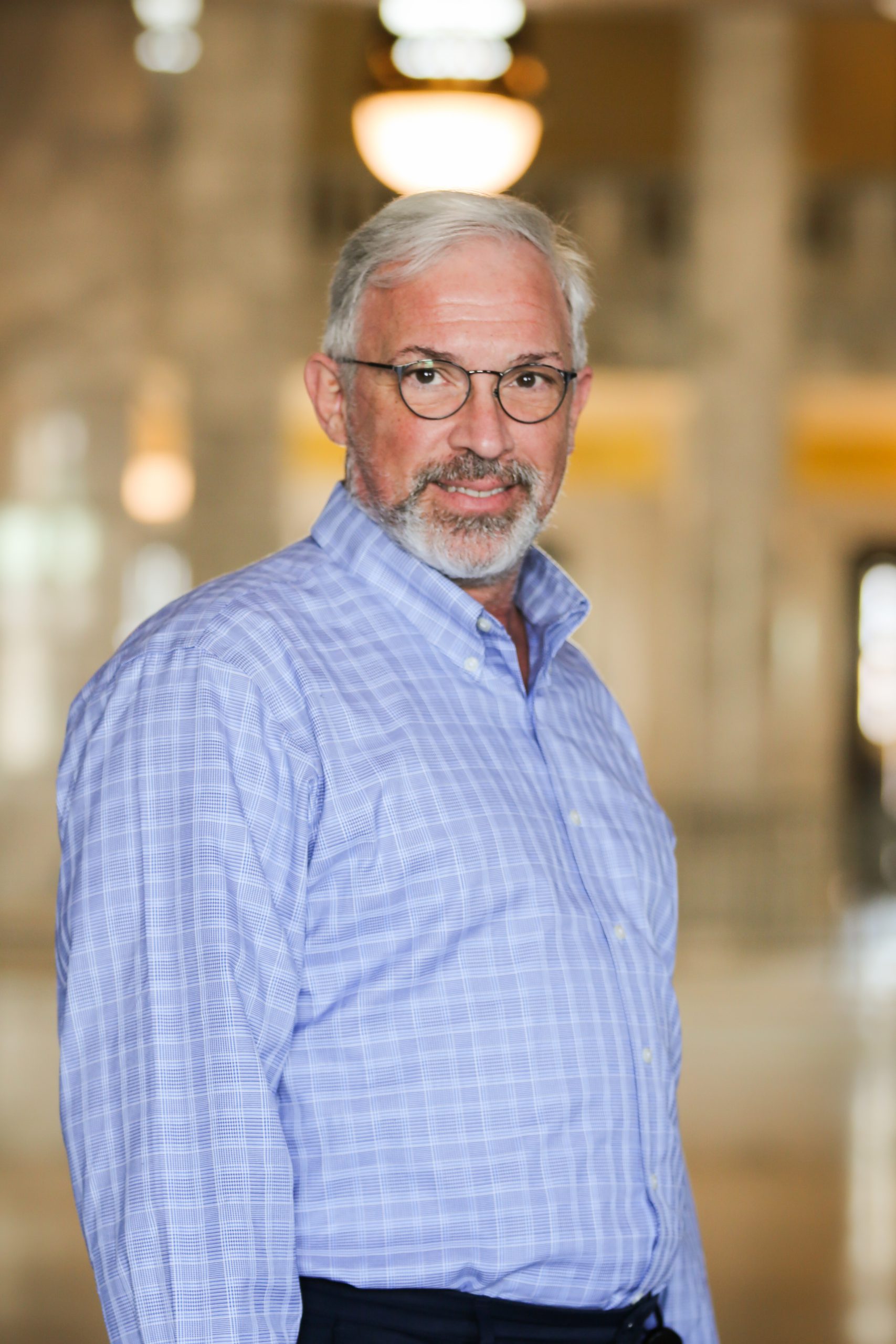 Older man with gray hair and beard, wearing glasses and a blue plaid shirt, indoors.