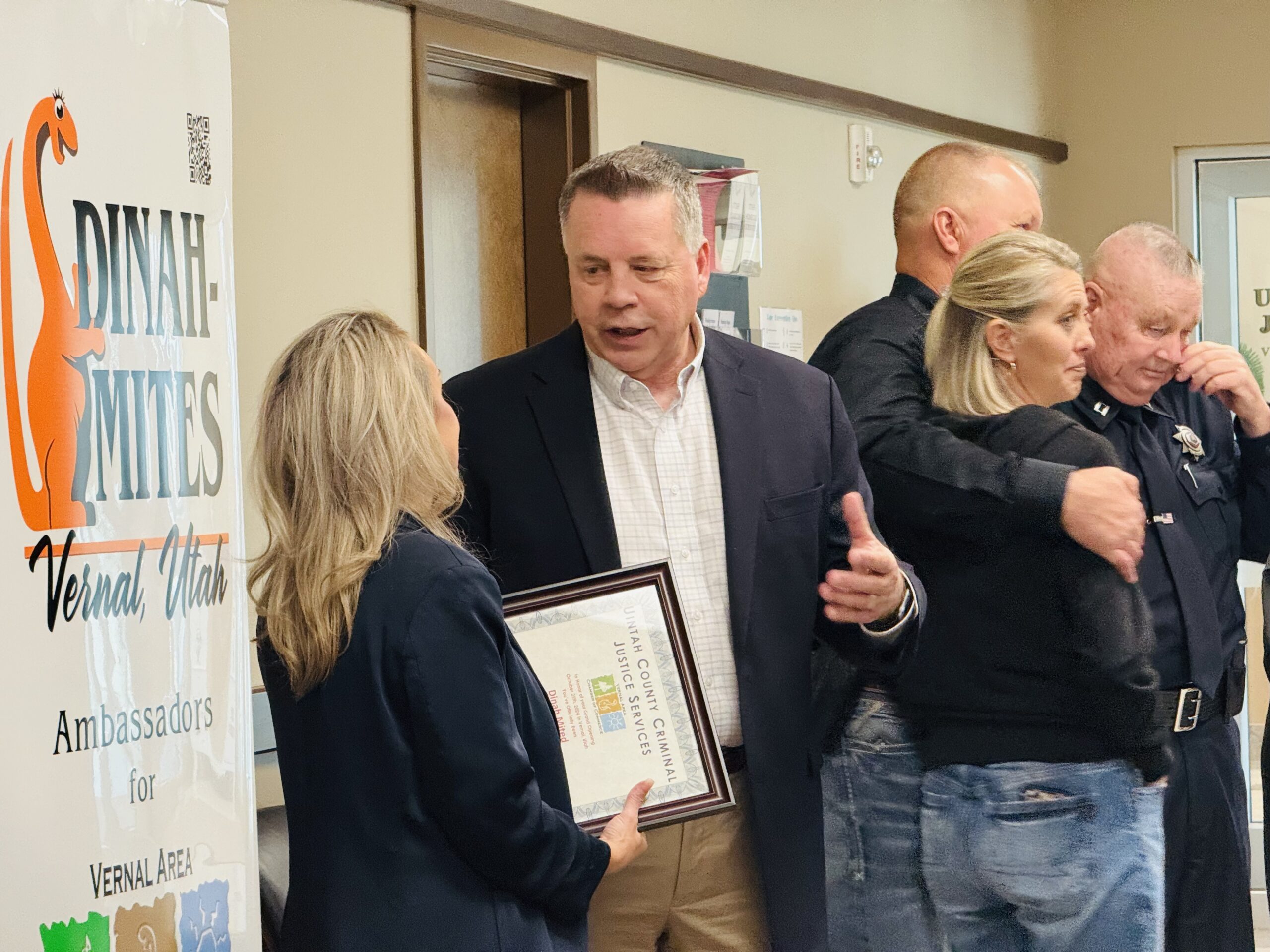 Two people talk as one holds a certificate; others interact in the background near a Dinah-Mites sign.