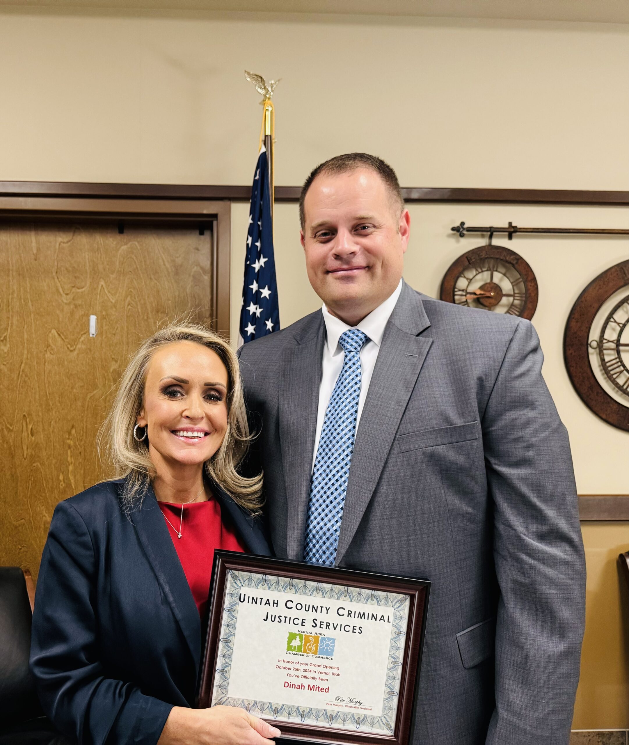 Two people in business attire hold a framed award in an office with flags and clocks.