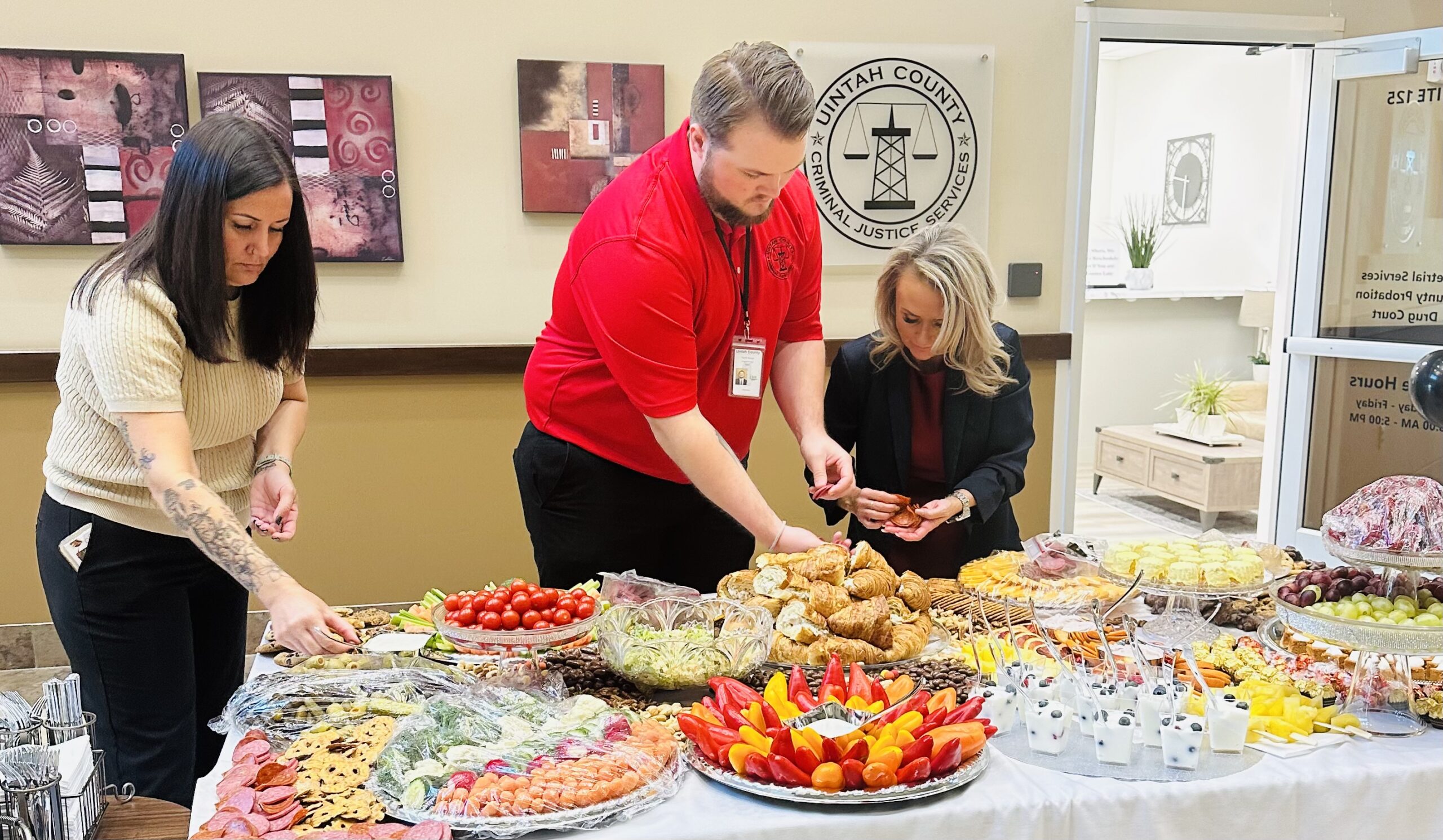 Three people prepare a large table of assorted snacks and food at an indoor event.