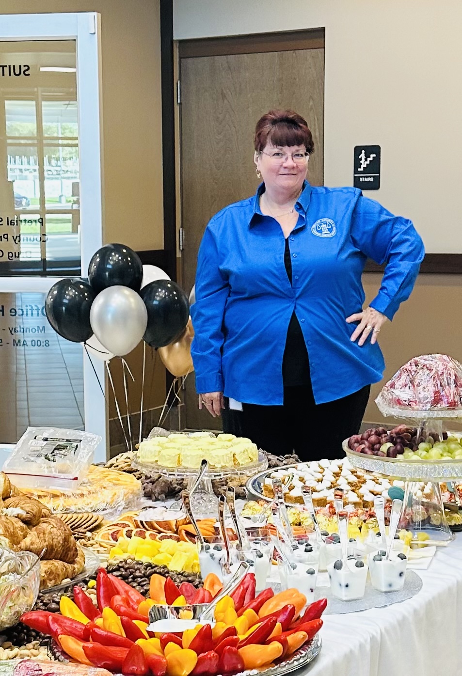 Woman in blue shirt stands by a table with assorted food, desserts, and black balloons.