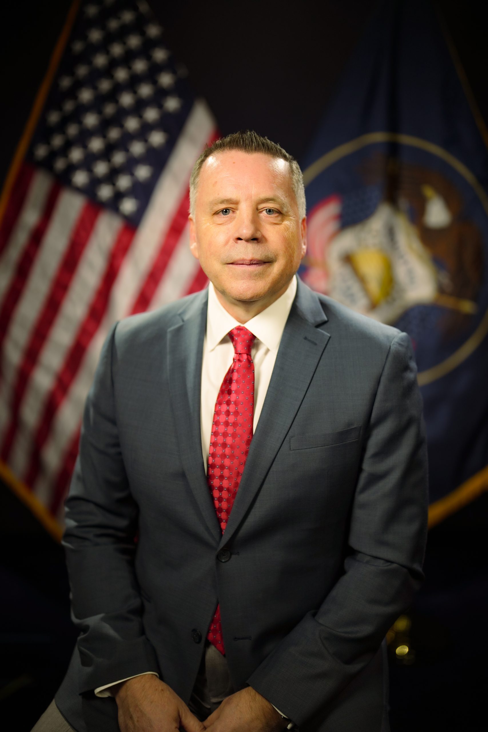 Man in suit and red tie sitting, with American and state flags in the background.