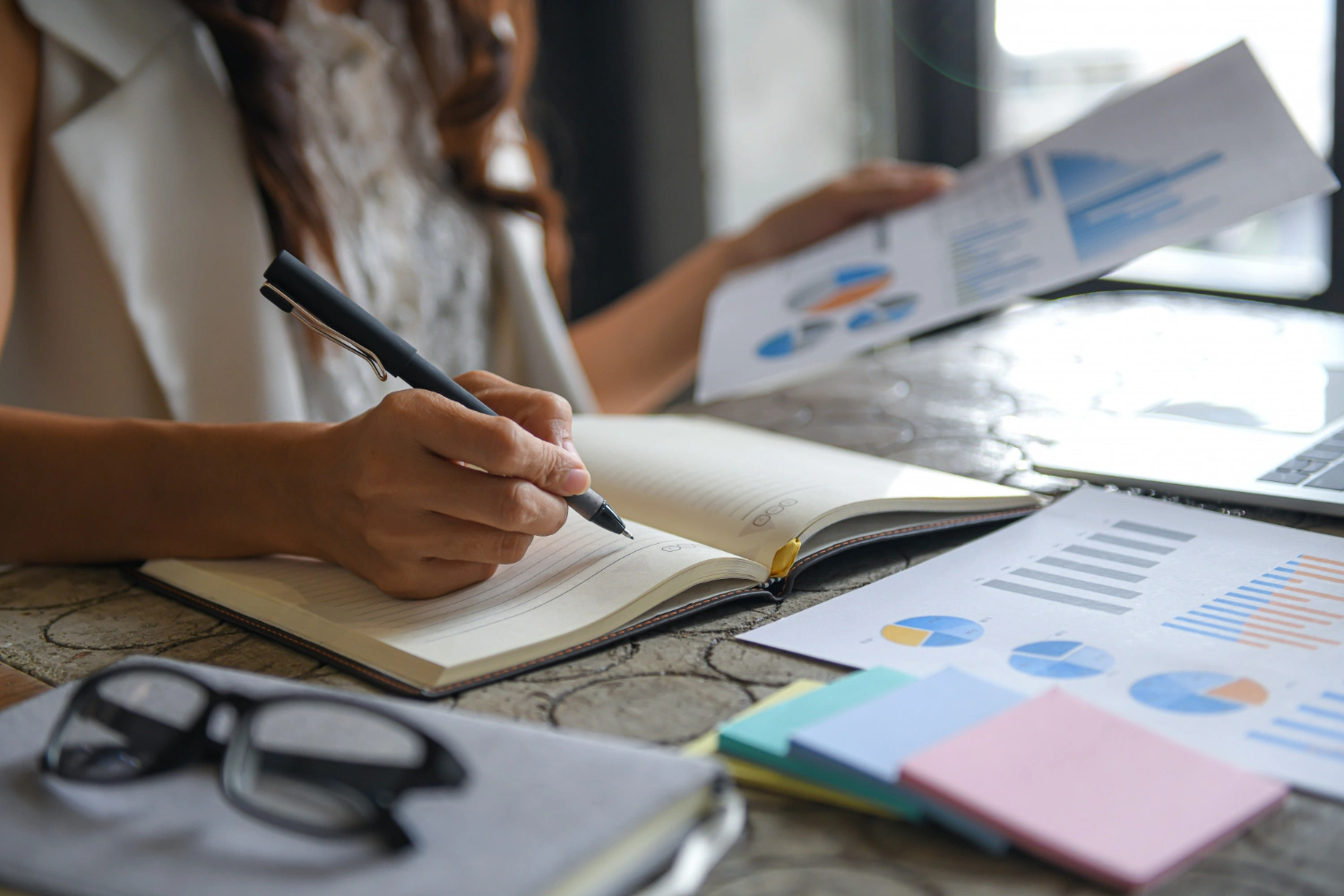 Person writing in a notebook with charts, graphs, and documents on the table.