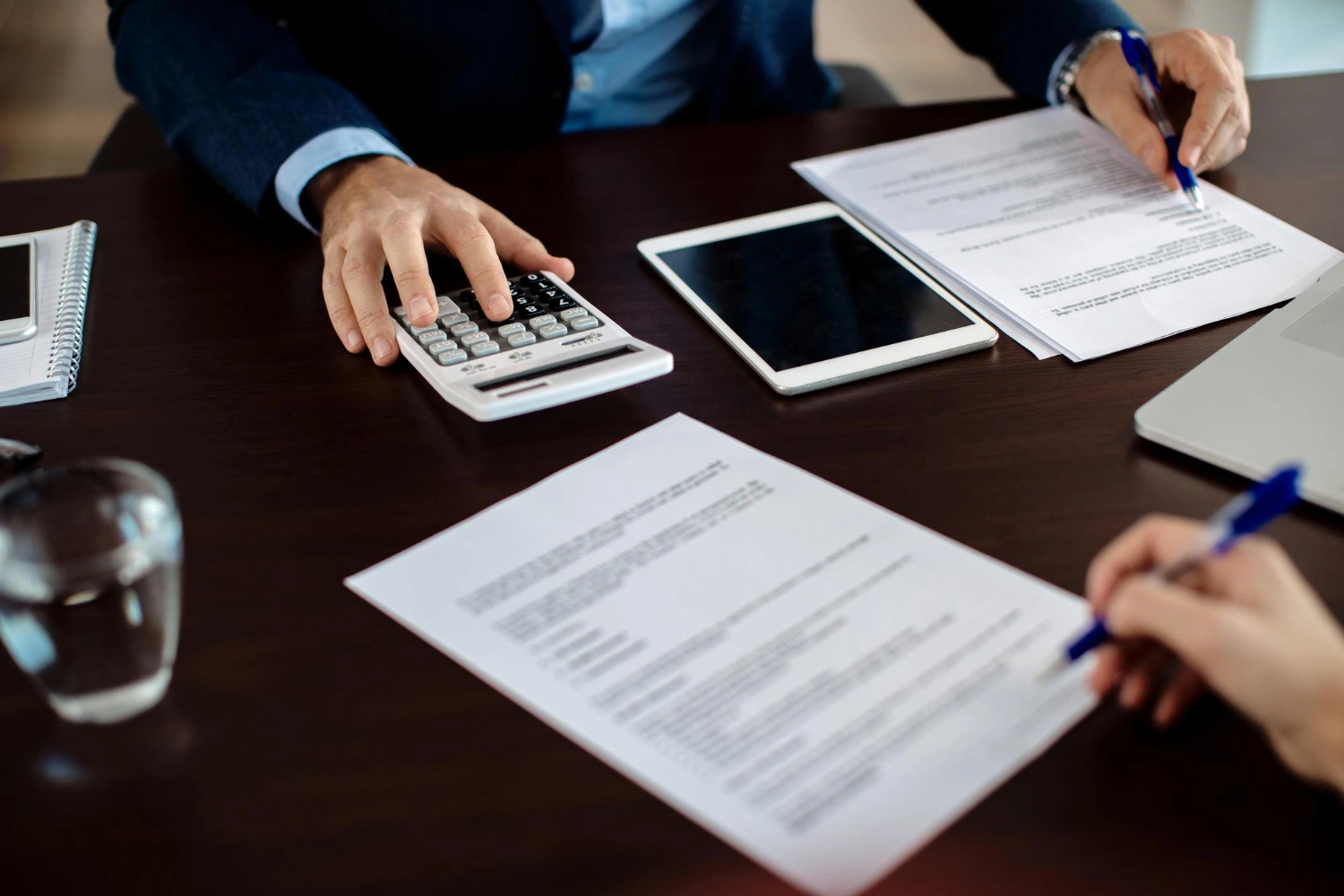 Two people work with documents, a calculator, and a tablet at a dark desk.