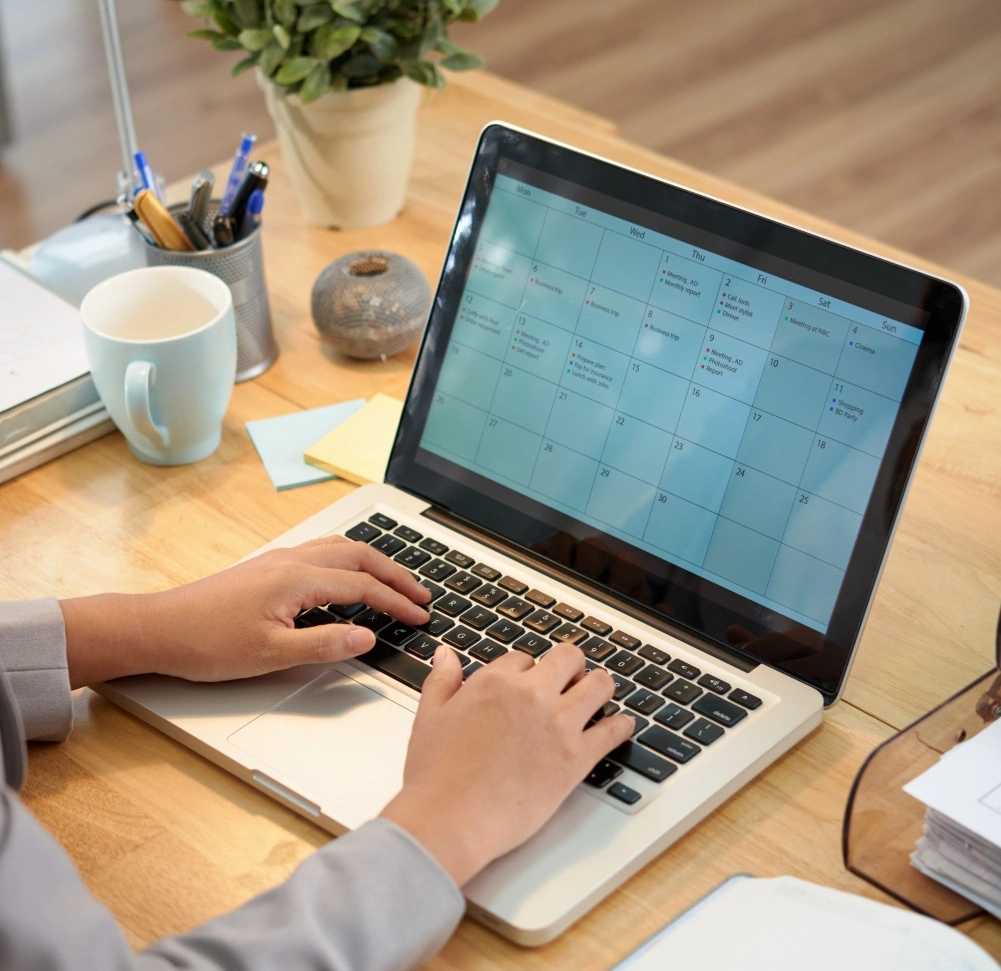 Person typing on a laptop displaying a calendar, with office supplies on the desk.