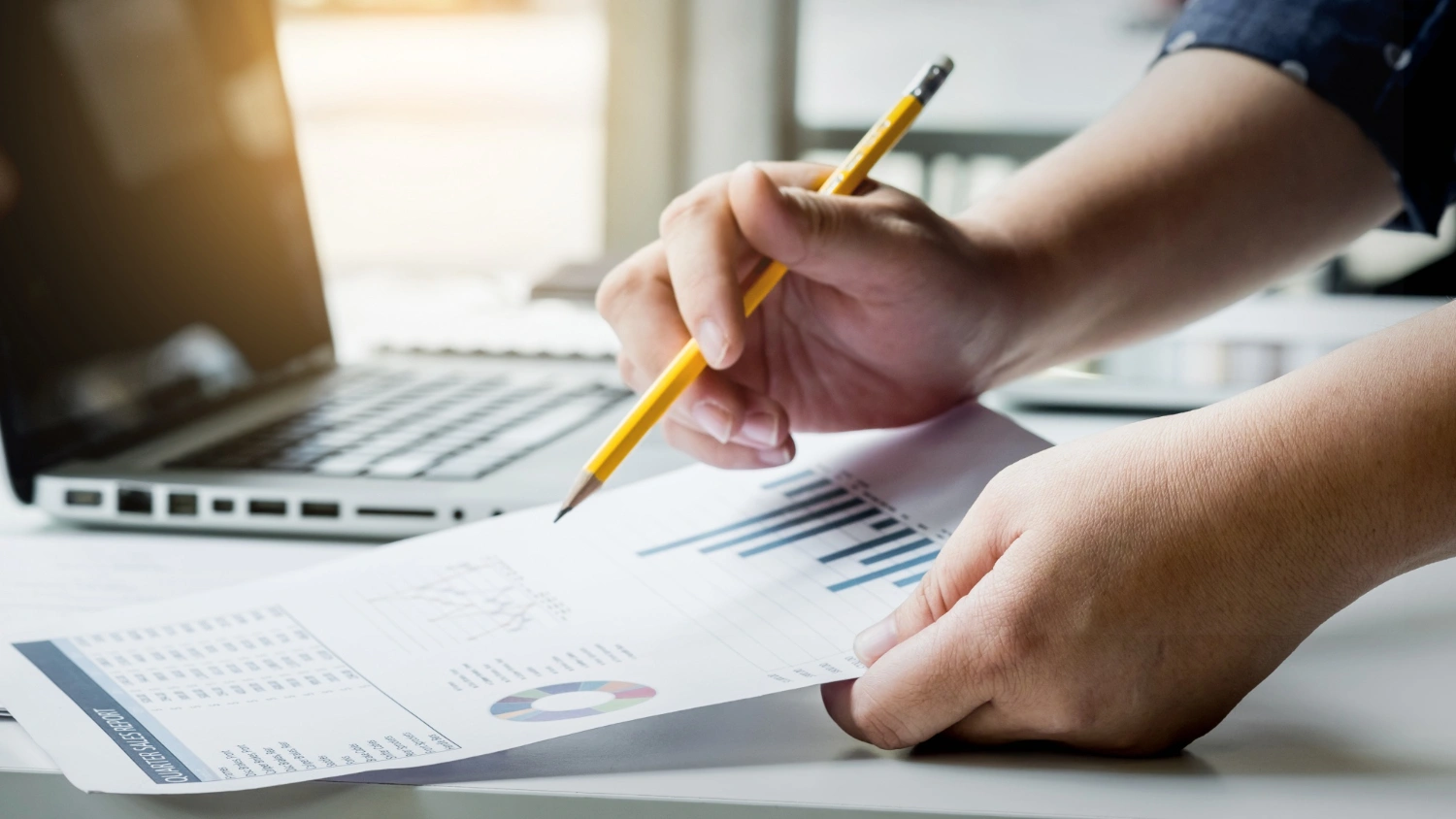 Person analyzing printed charts with a pencil next to an open laptop on a desk.