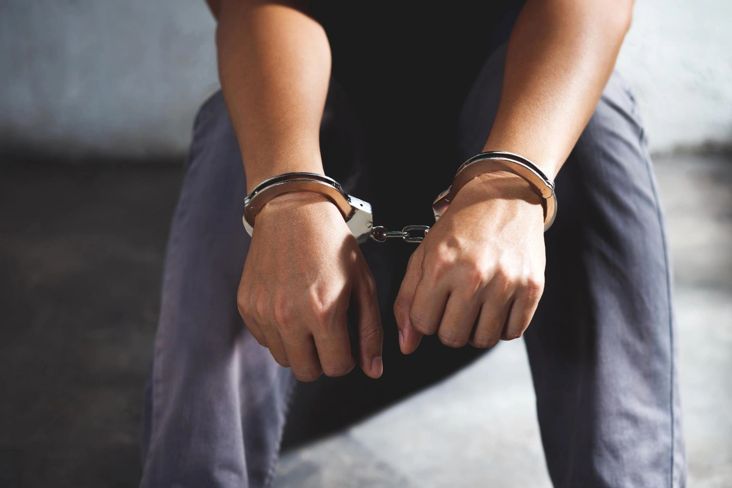 Person sitting with hands in metal handcuffs, wearing gray pants, indoors.