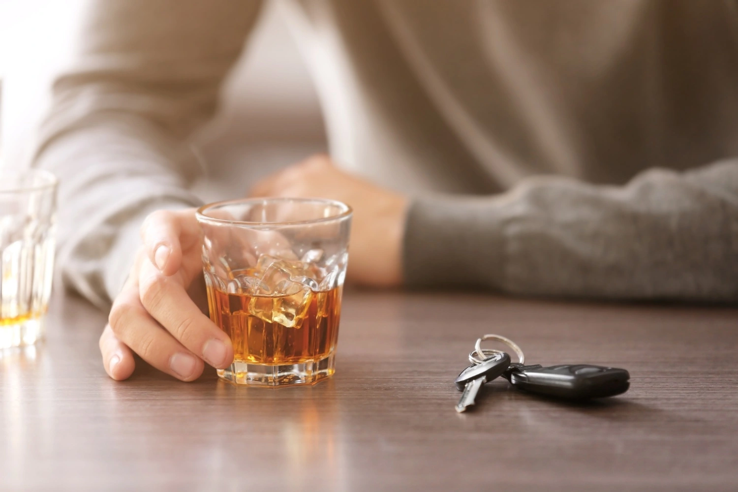 Person holding a glass of whiskey with car keys on the table nearby.