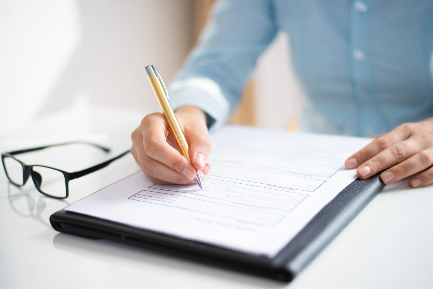 Person filling out a form on a clipboard with glasses on the desk nearby.