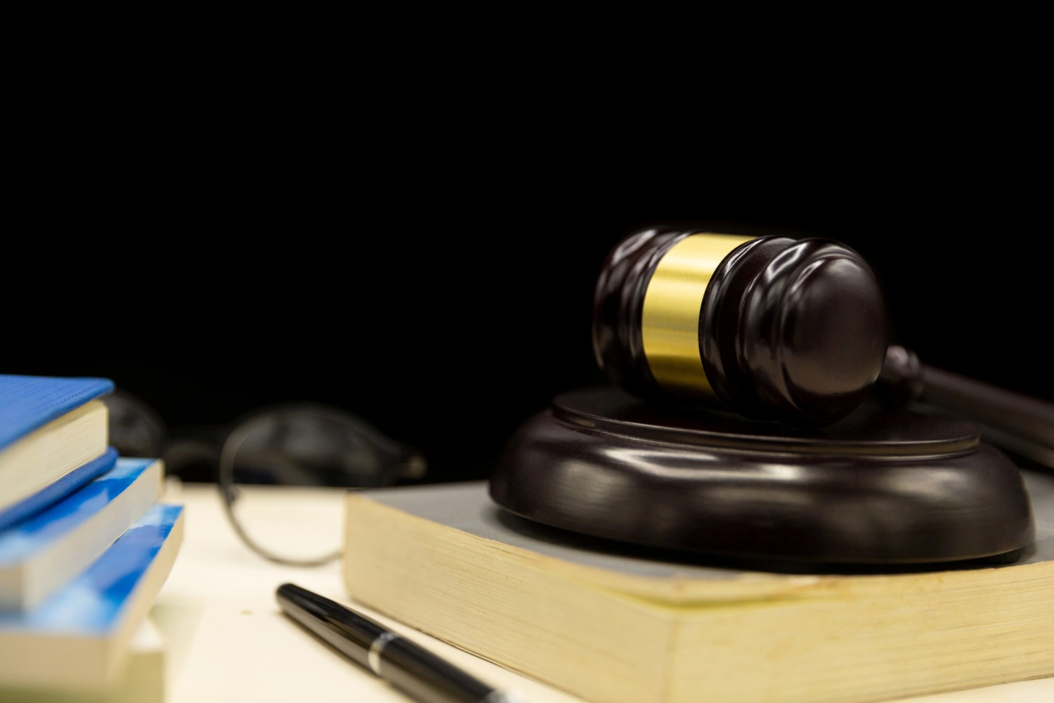 A judges gavel on a book, with a pen and stacked books nearby, against a black background.