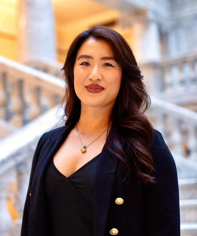Woman with long dark hair in a black outfit standing in front of marble stairs indoors.