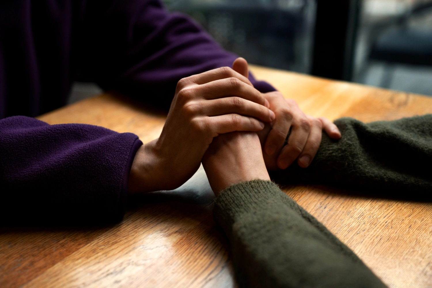 Two people holding hands across a wooden table, offering comfort and support.