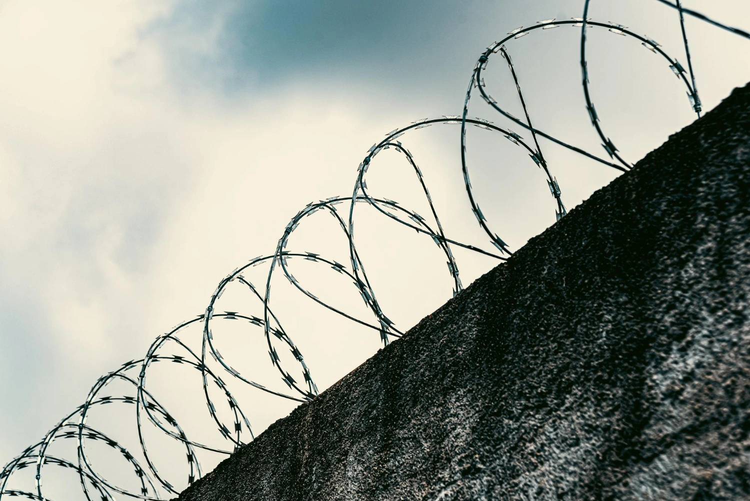 Barbed wire spirals atop a concrete wall against a cloudy sky.
