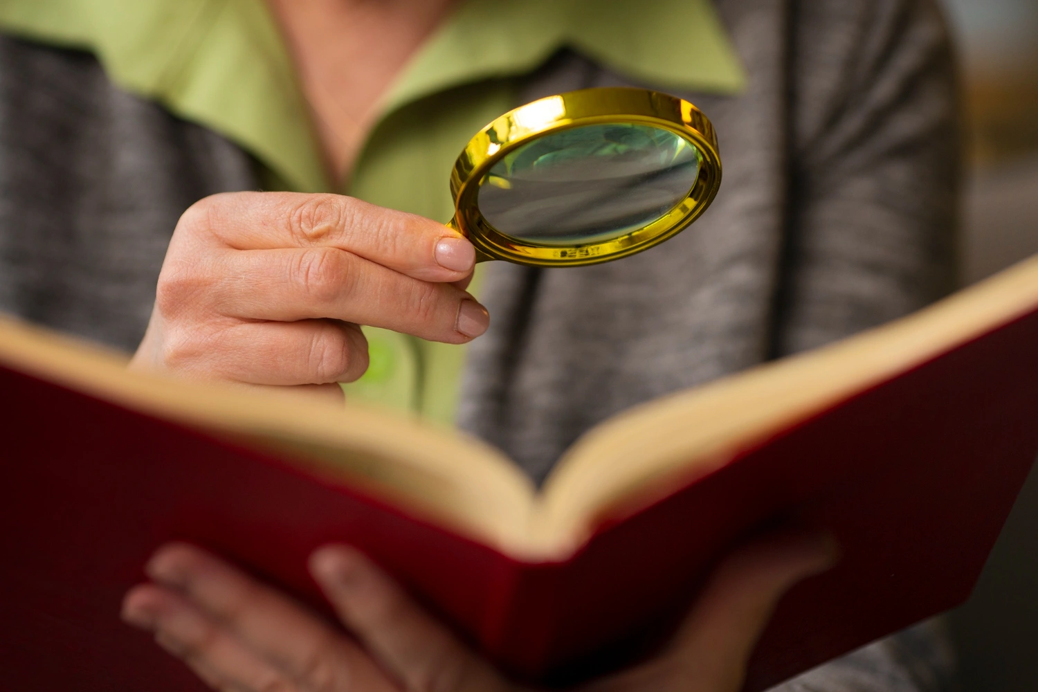 Person holding a magnifying glass over an open book with a red cover.