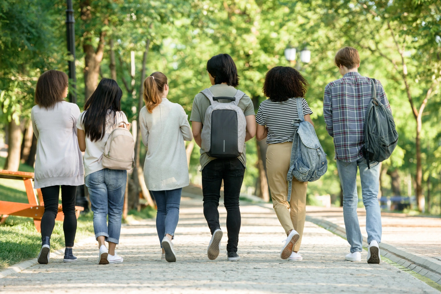 Six young people walk together down a park path with backpacks on a sunny day.